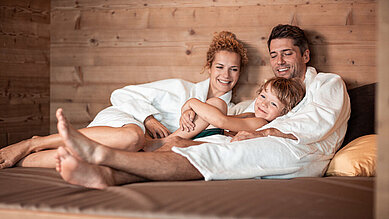 Familie mit Kind liegt gemütlich und entspannt im Bademantel auf einer Liege im Wellnessbereich des Hotels Family Home Alpenhof.